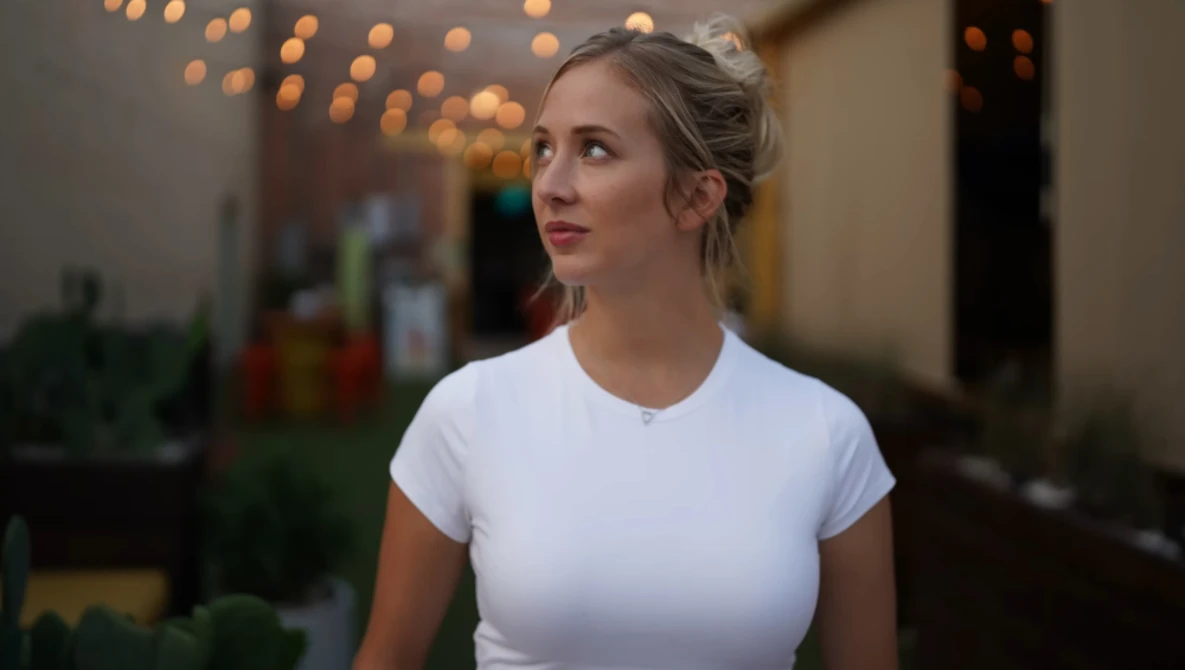 Woman in white t-shirt looking upward in indoor setting with warm bokeh lighting.