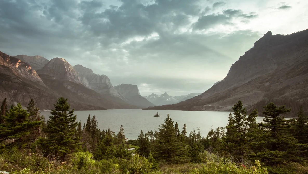 Mountain lake surrounded by evergreen forest with dramatic peaks and storm clouds overhead.