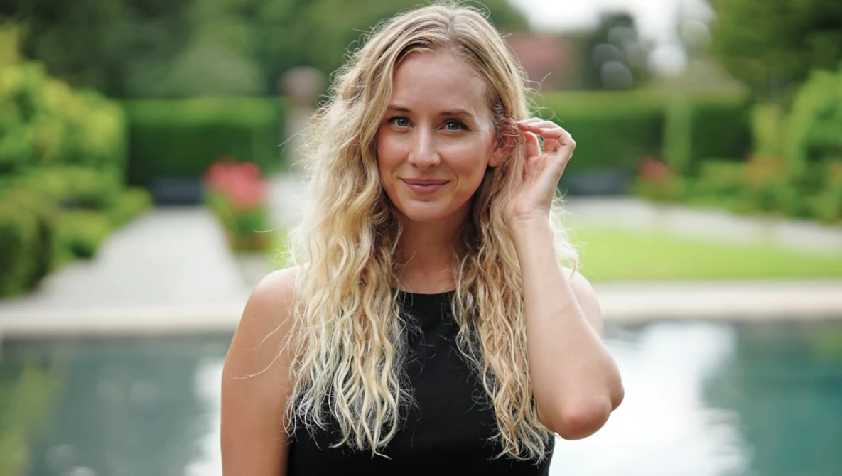 Blonde woman in black sleeveless top posing outdoors by a pool with manicured gardens in background.