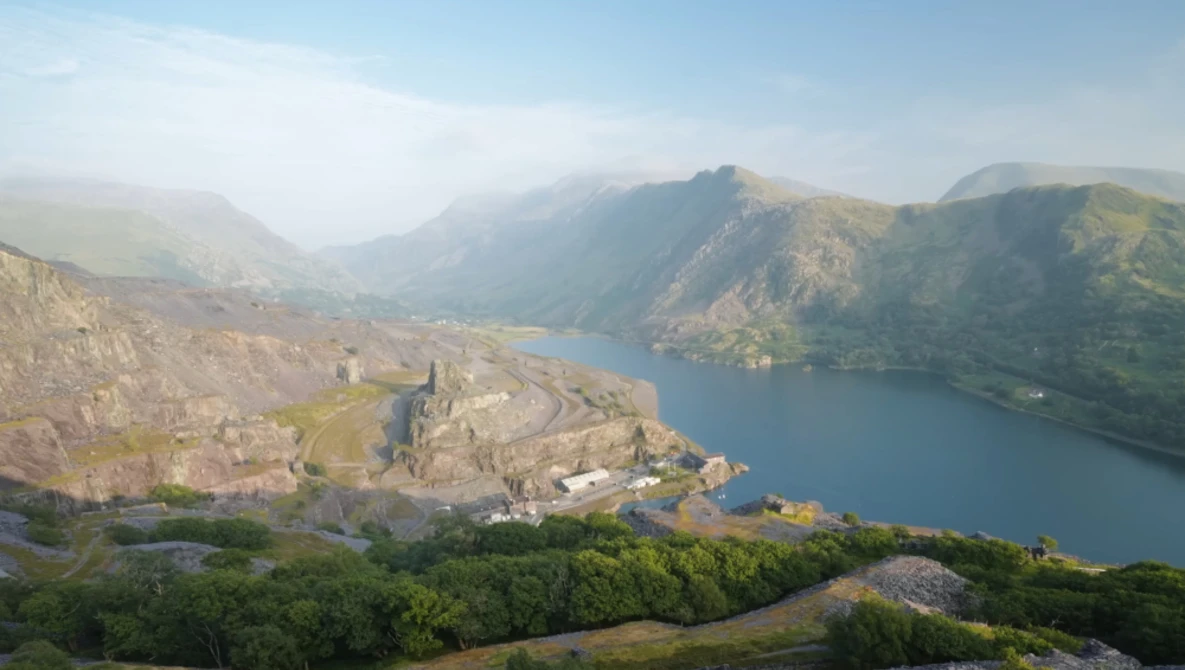 Medieval castle ruins on a rocky peninsula surrounded by a deep blue lake, with forested mountains in the background under hazy morning light.