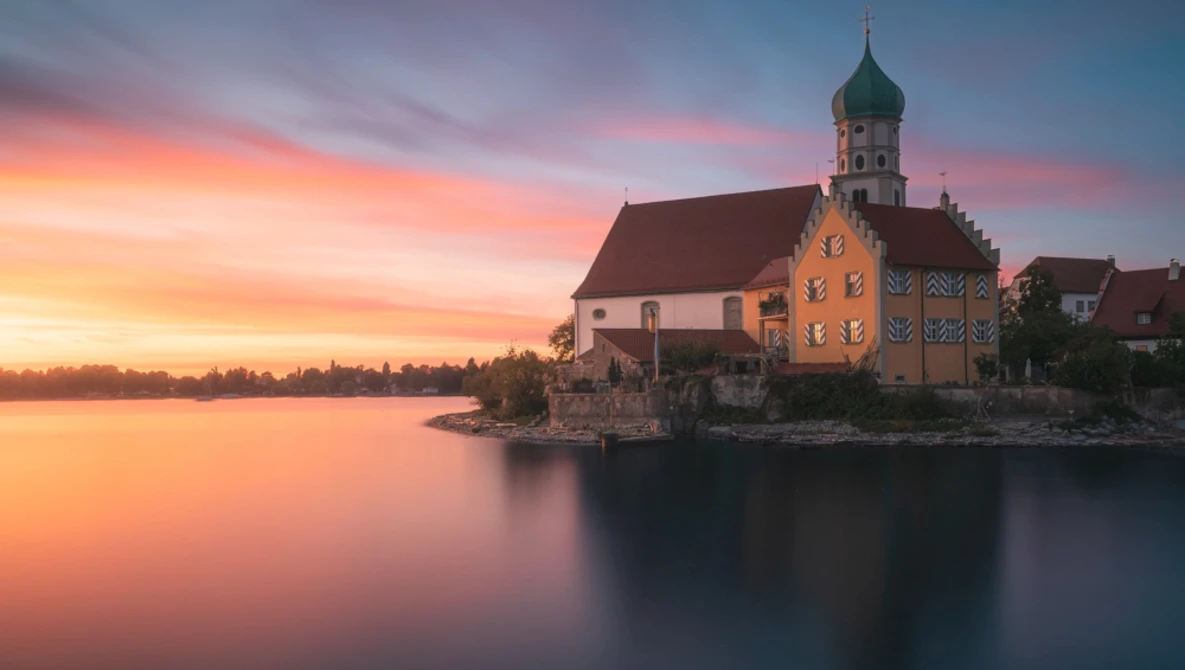 Waterfront church with green spire reflected in still lake at sunset.
