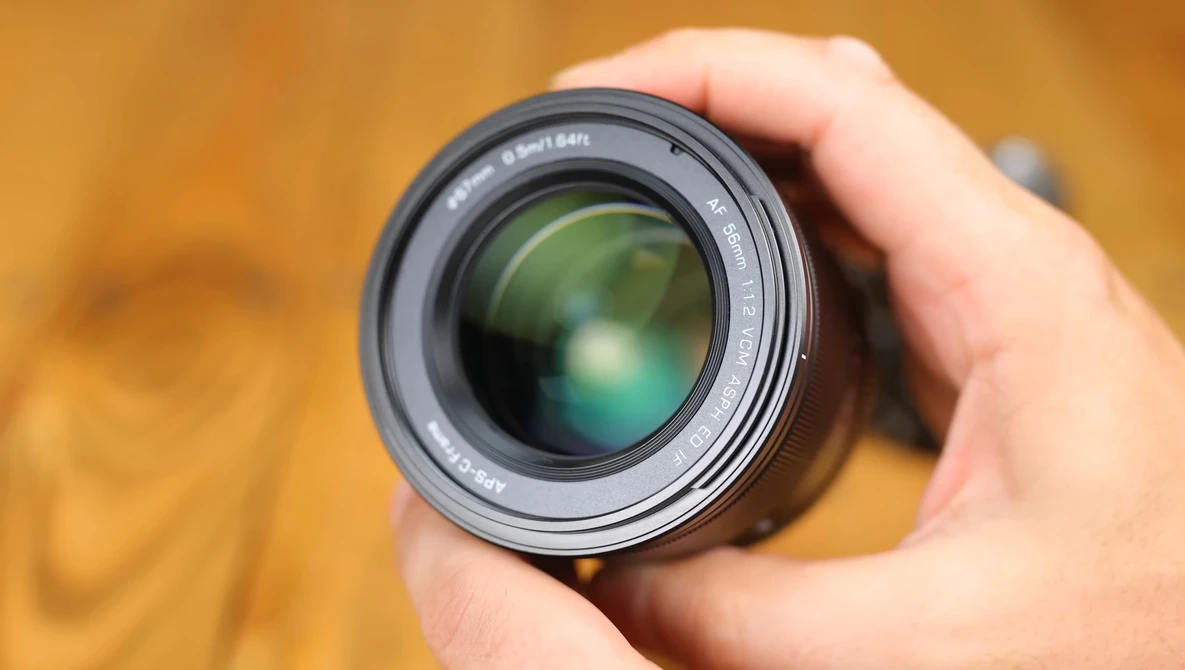 Camera lens held in hand against warm, blurred background. Glass elements visible through front optic.