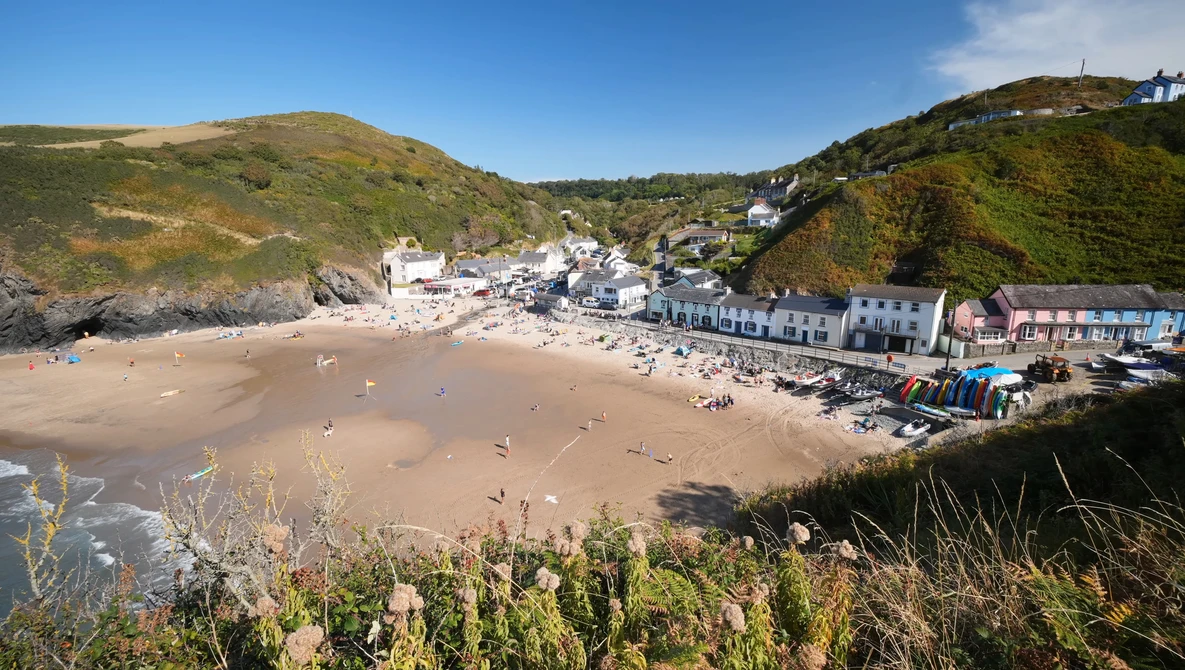 Coastal beach village nestled between green hillsides with sandy beach and beachfront buildings in the foreground.