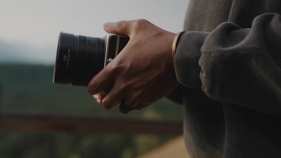 Photographer holding a telephoto lens while looking toward a blurred green landscape in overcast daylight.
