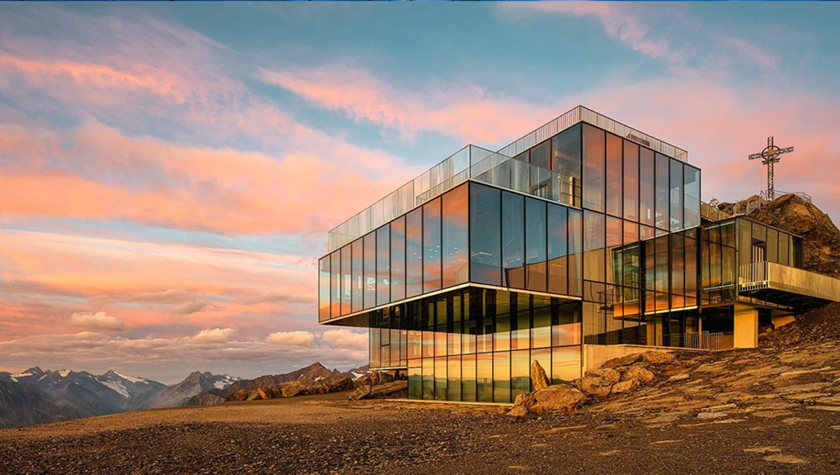 Modern glass and steel building with cantilever design perched on rocky alpine terrain at sunset.