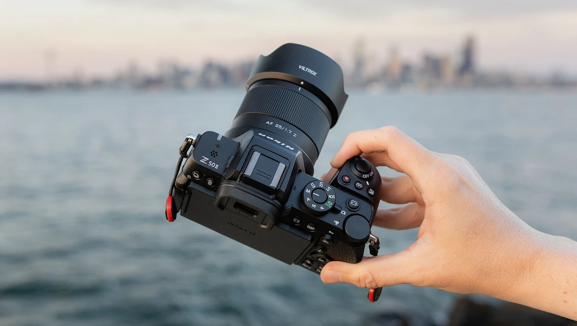 Hand holding a mirrorless camera with attached telephoto lens over water with city skyline in background.