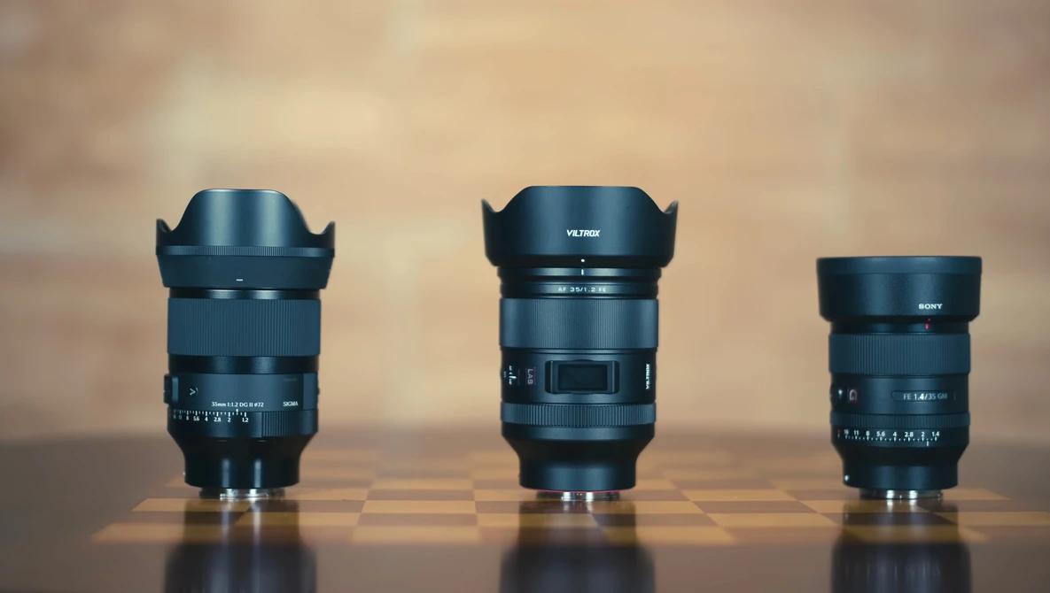 Three camera lenses of varying sizes arranged side by side on a reflective surface against a warm, blurred background.