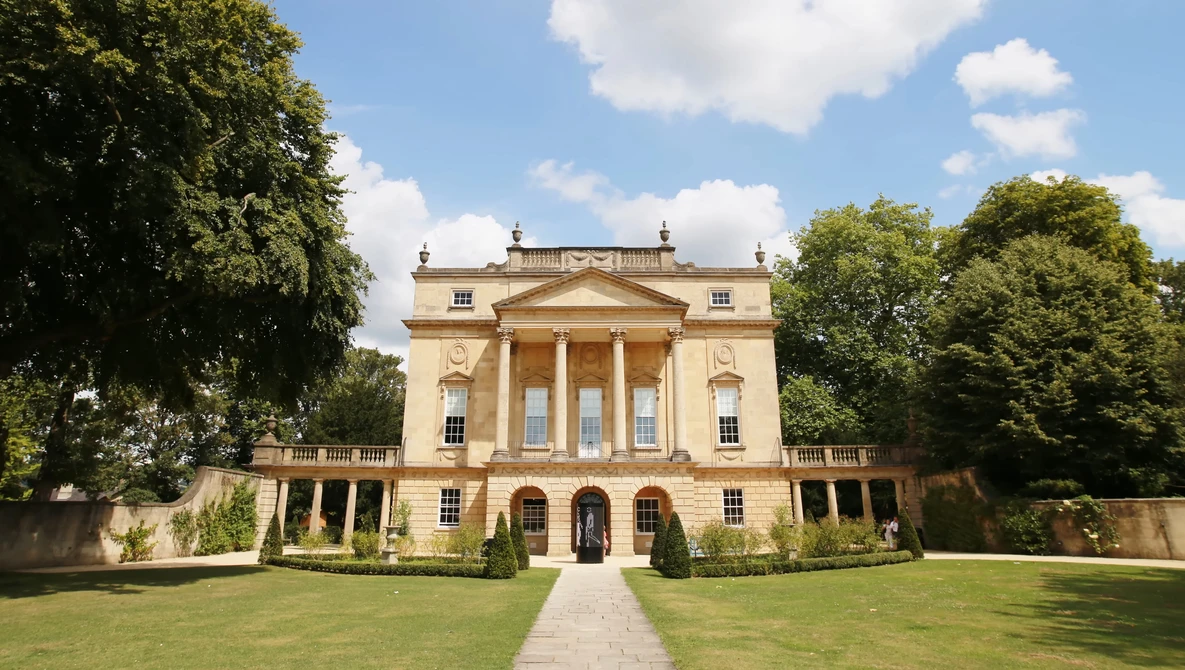 Neoclassical stone mansion with columned portico, symmetrical facade, and manicured grounds under blue sky.