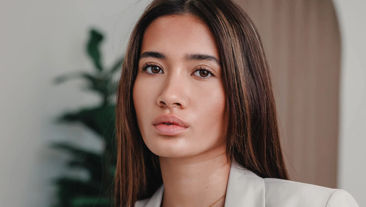 Woman with long dark hair wearing a white blazer, gazing directly at camera with neutral expression.
