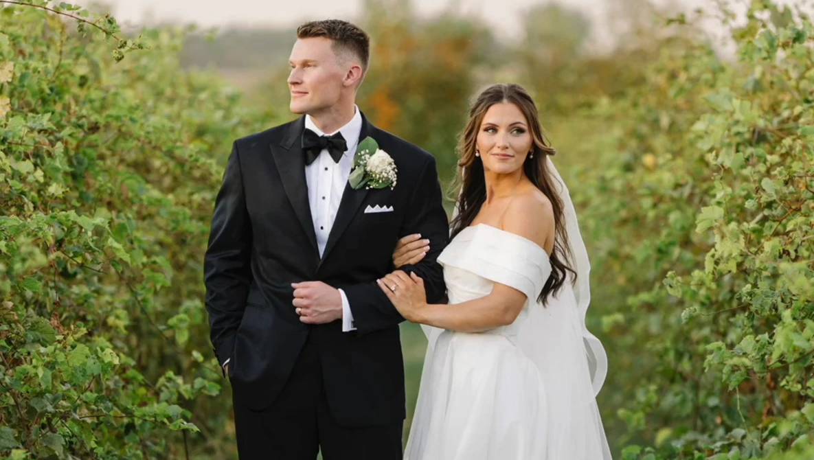 Bride and groom walking together through a vineyard in formal wedding attire.