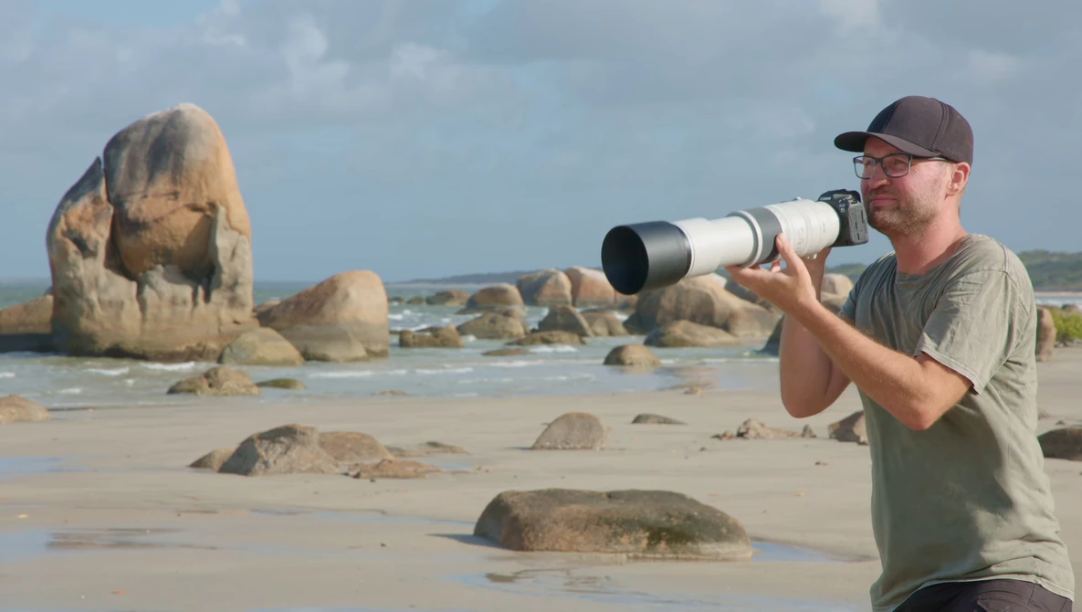Photographer holding a telephoto lens on a beach with large granite rock formations.