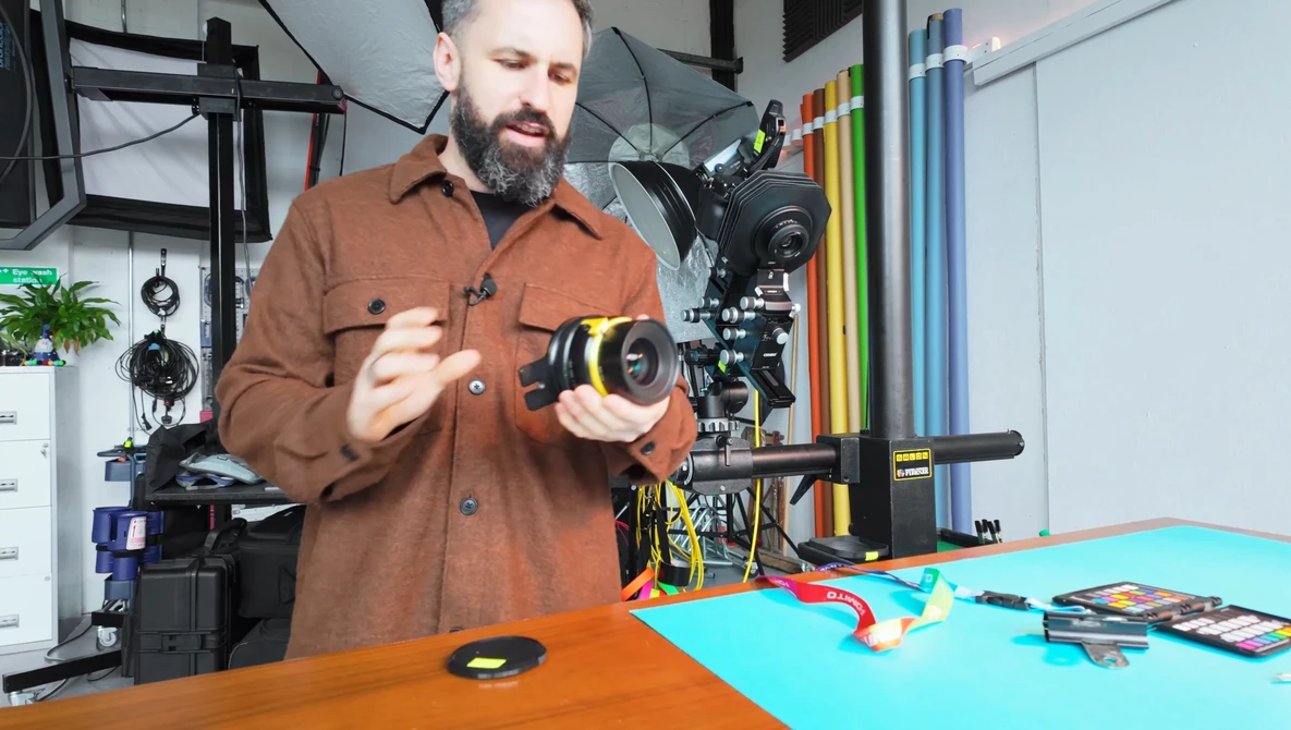 Bearded man in brown shirt holding a cinema camera lens in a studio workspace.