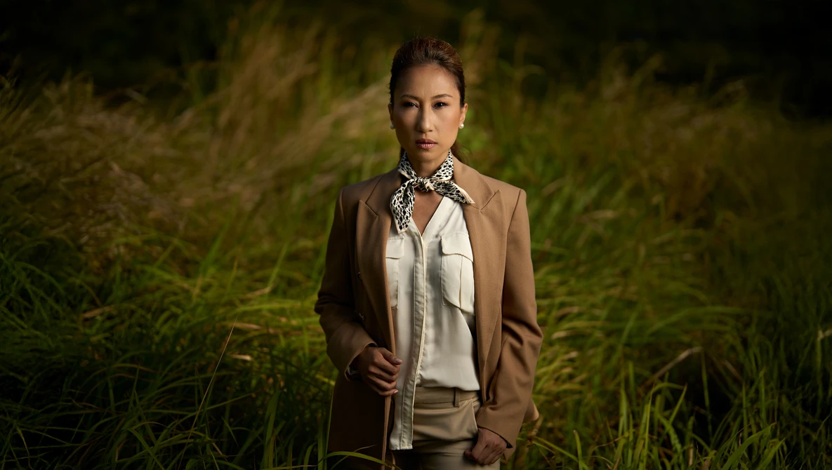 Woman in tan blazer and patterned scarf standing in tall grass at dusk.