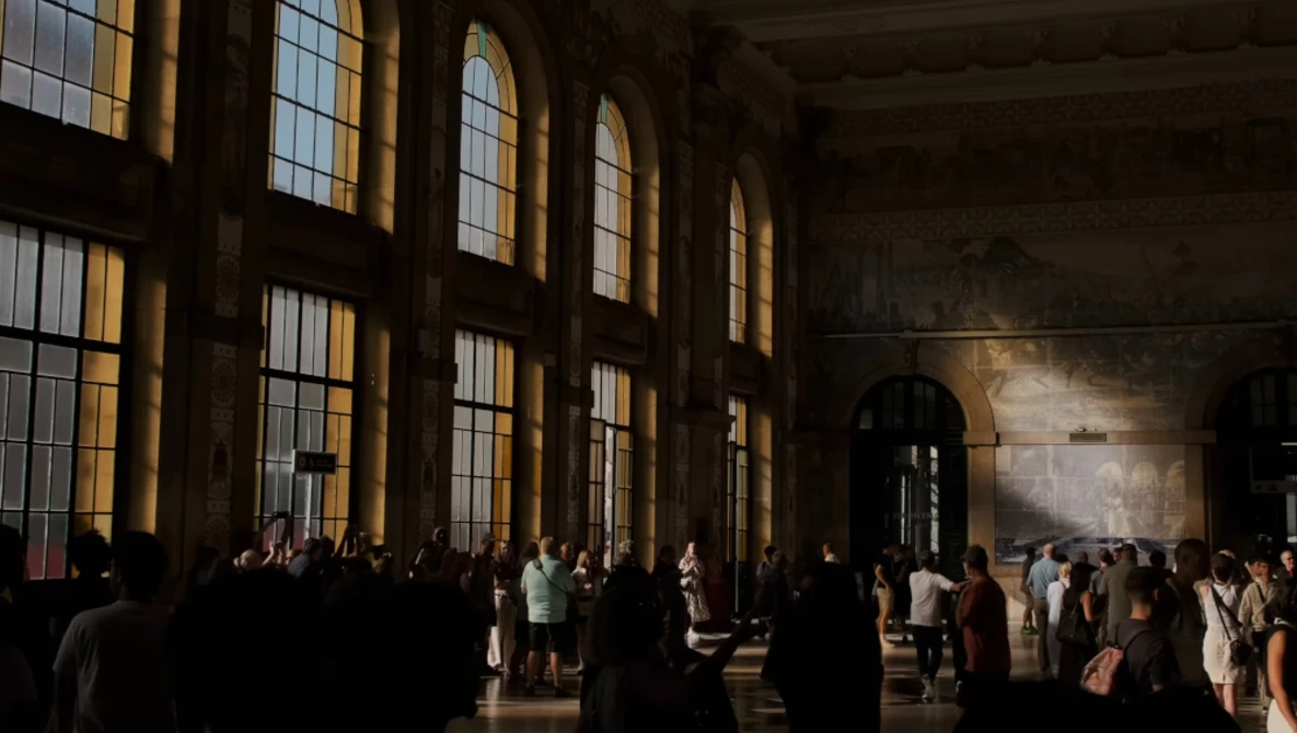 Crowds of visitors walking through a grand hall with tall arched windows casting golden light across the space.