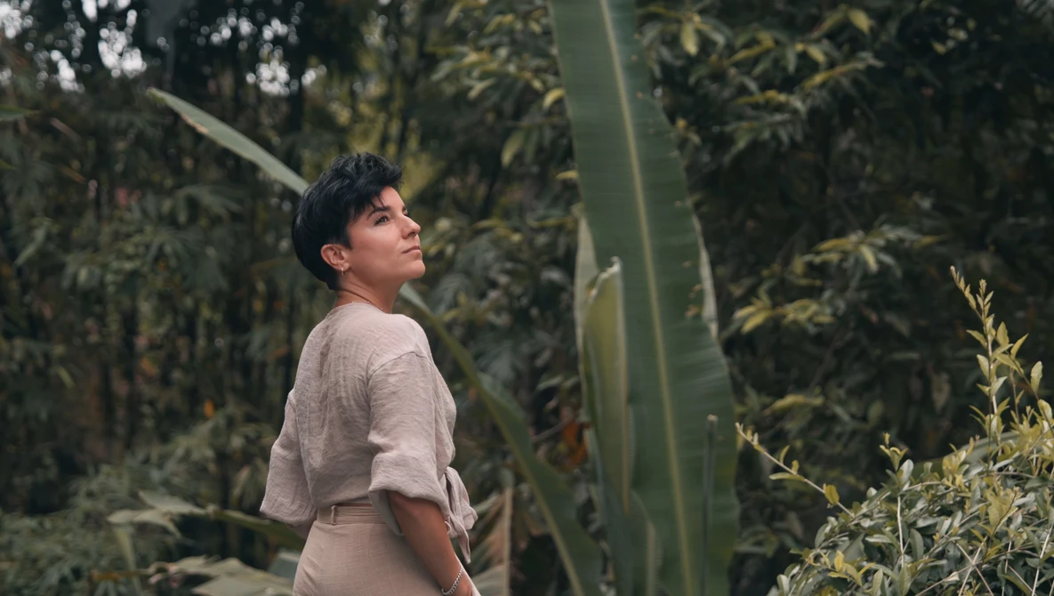 Person in beige shirt standing among tall green plants and cacti in a greenhouse environment.