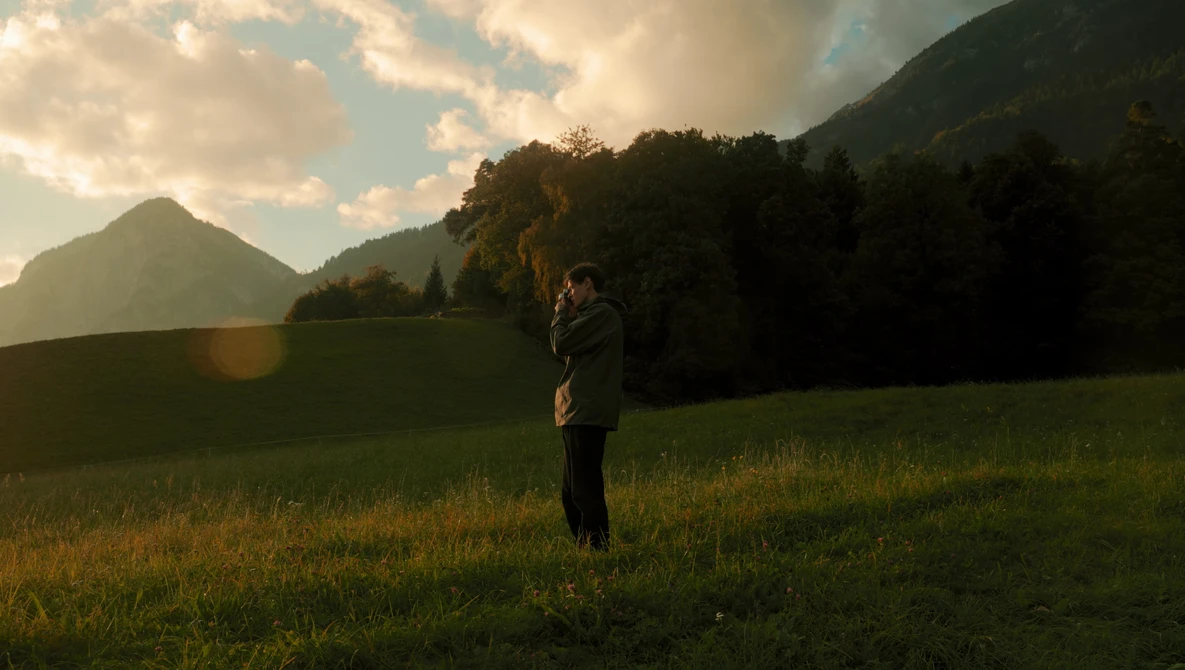 Person standing in a grassy meadow with forested mountains in the background during golden hour.