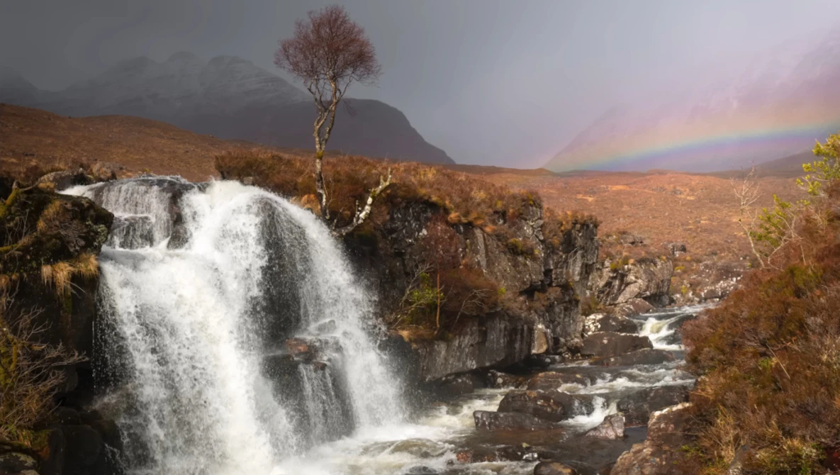 Waterfall cascading over dark rocks with rainbow arcing across misty valley landscape.