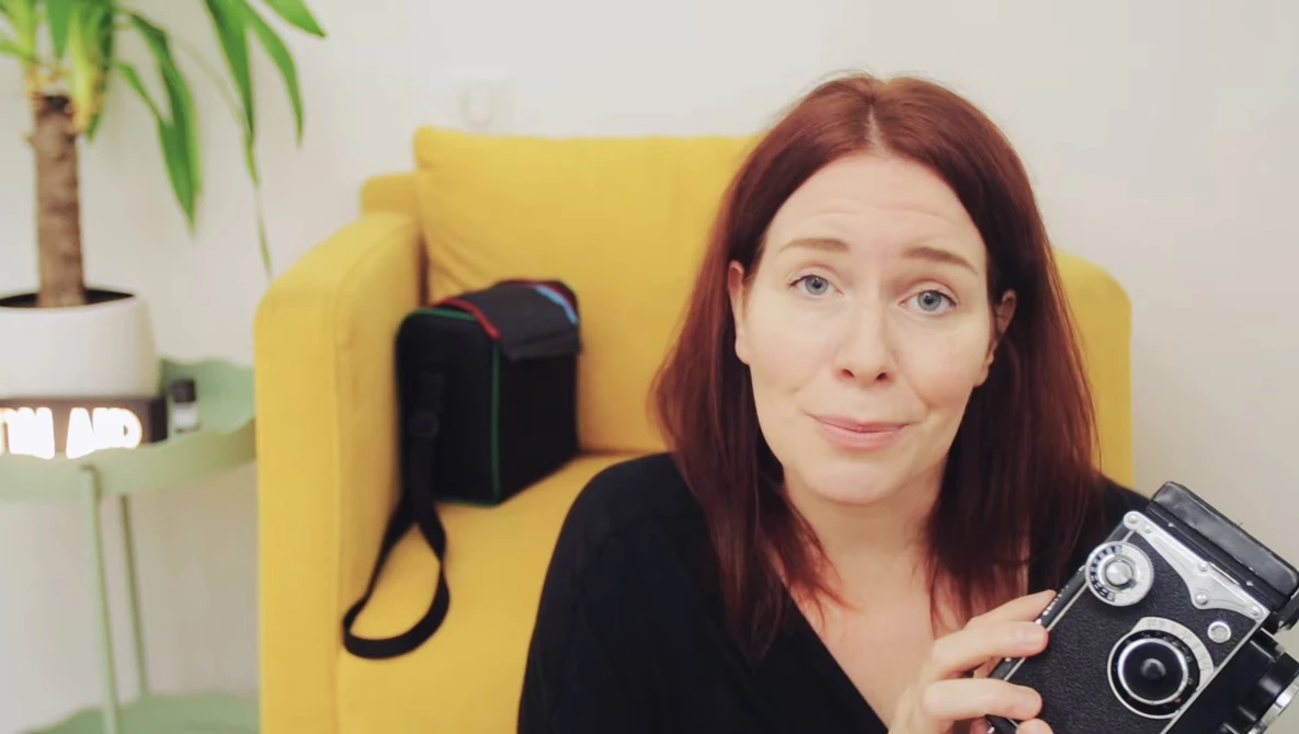 Woman with reddish-brown hair holding a vintage twin-lens reflex camera, seated in a yellow chair.