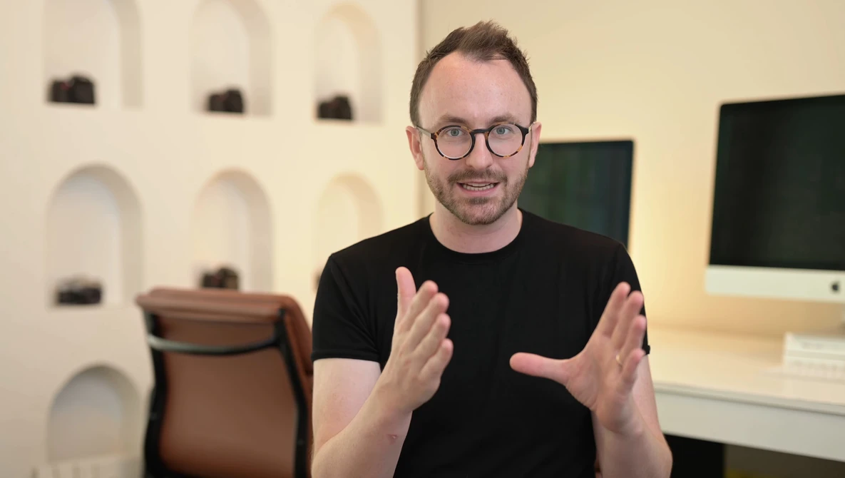 Man in black t-shirt and glasses gesturing while speaking in a modern studio setting.