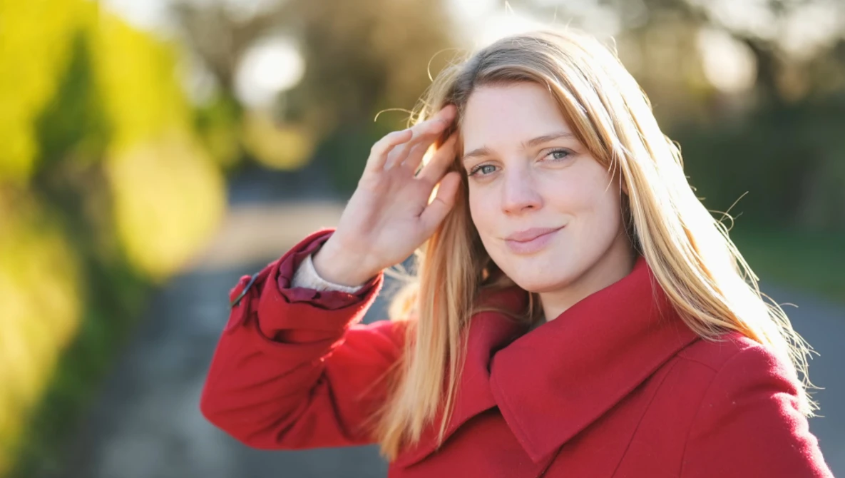 Woman with long strawberry blonde hair wearing a red coat, touching her hair and looking at camera outdoors.