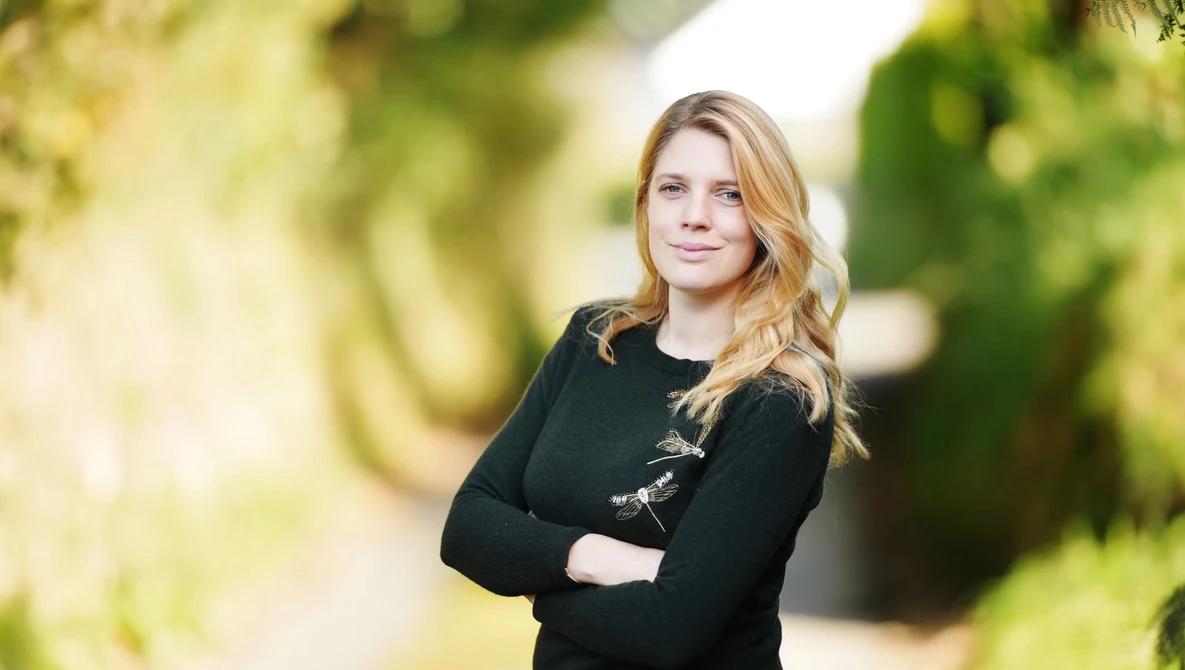 Woman with blonde wavy hair wearing a black cardigan with decorative clasps, standing with crossed arms in an outdoor garden setting.