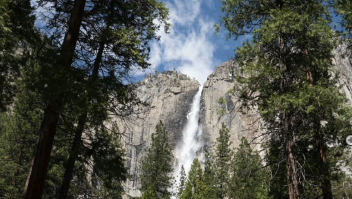 Waterfall cascading down granite cliffs framed by towering evergreen trees and blue sky.