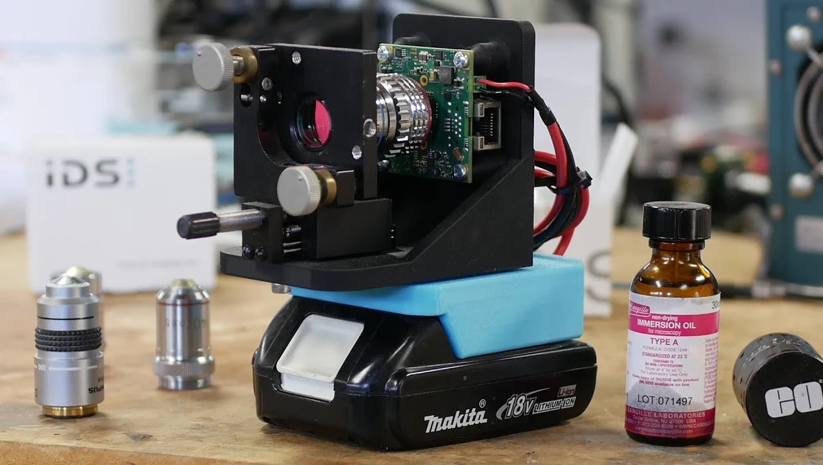 DIY optical microscope setup built with a Makita battery, circuit board, and lens components on a workbench.