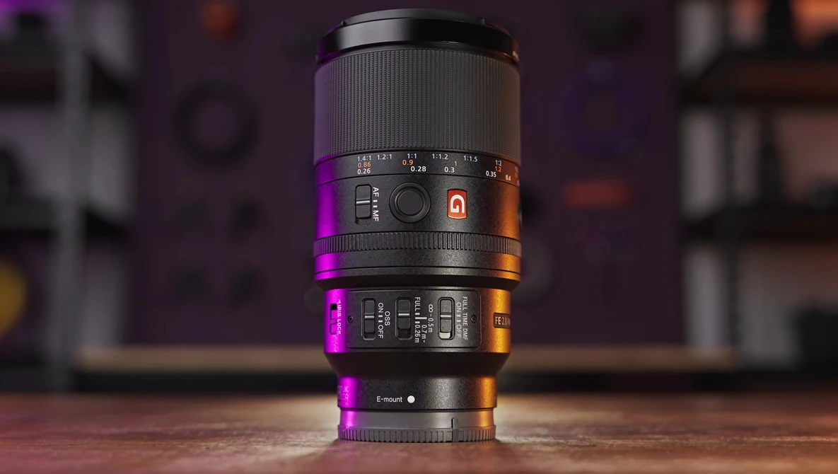 Camera lens with focus ring and aperture markings, lit by magenta and orange accent lighting against blurred background.