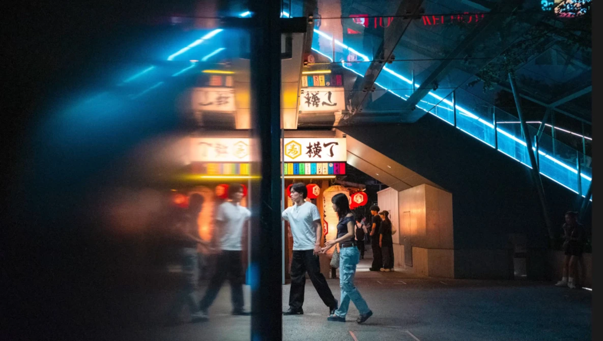 Night street scene with neon signage and blurred figures walking past storefronts.