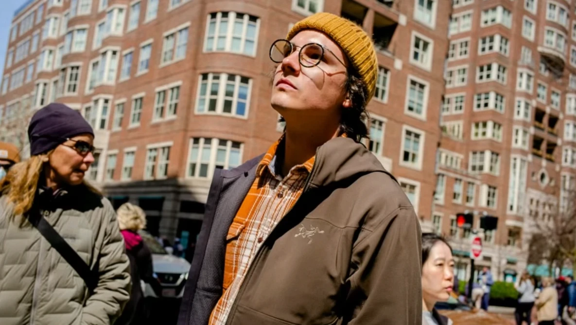 Person wearing glasses and yellow beanie looking upward on a city street with brick buildings behind.