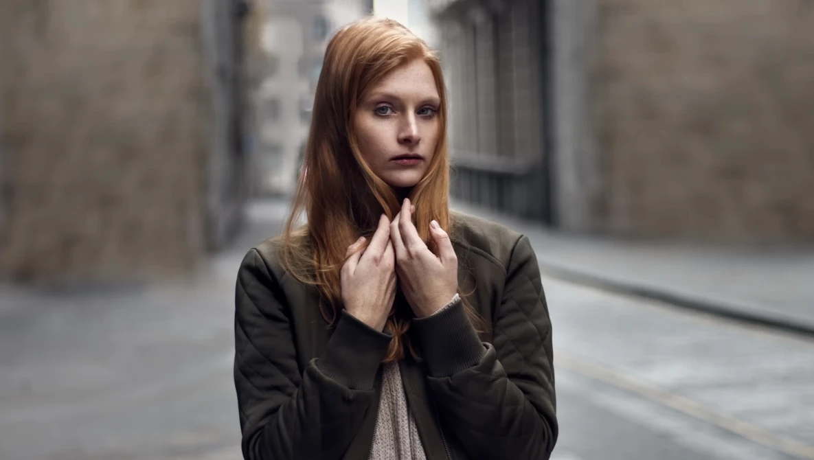 Woman with reddish-brown hair wearing a black jacket on an urban street, touching her neck while looking toward the camera.