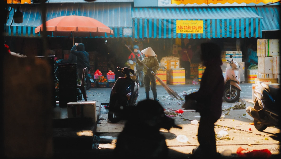 Street market scene viewed through silhouetted foreground figures, with vendors and blue corrugated metal structures under warm daylight.