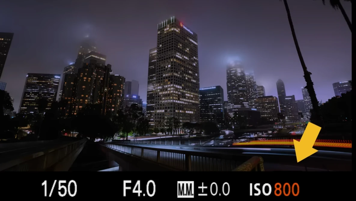 Urban skyline at dusk with illuminated skyscrapers reflected in still water, with camera settings displayed as an overlay.