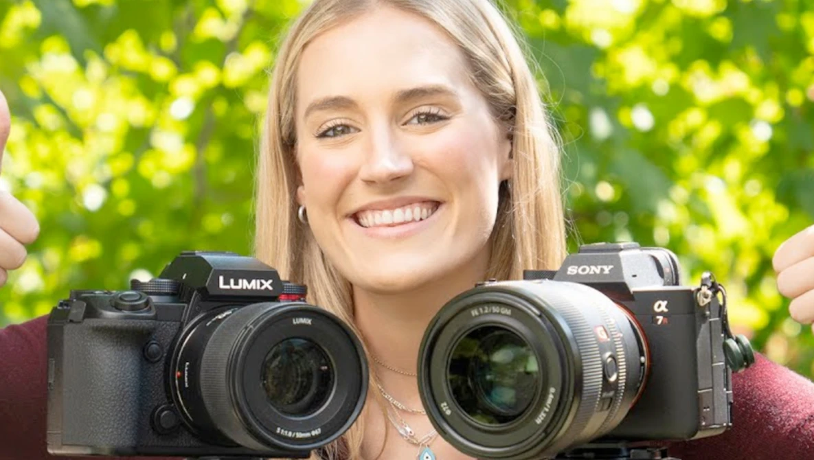 Woman holding a Lumix mirrorless camera in one hand and a Sony Alpha mirrorless camera in the other against a bright green bokeh background.