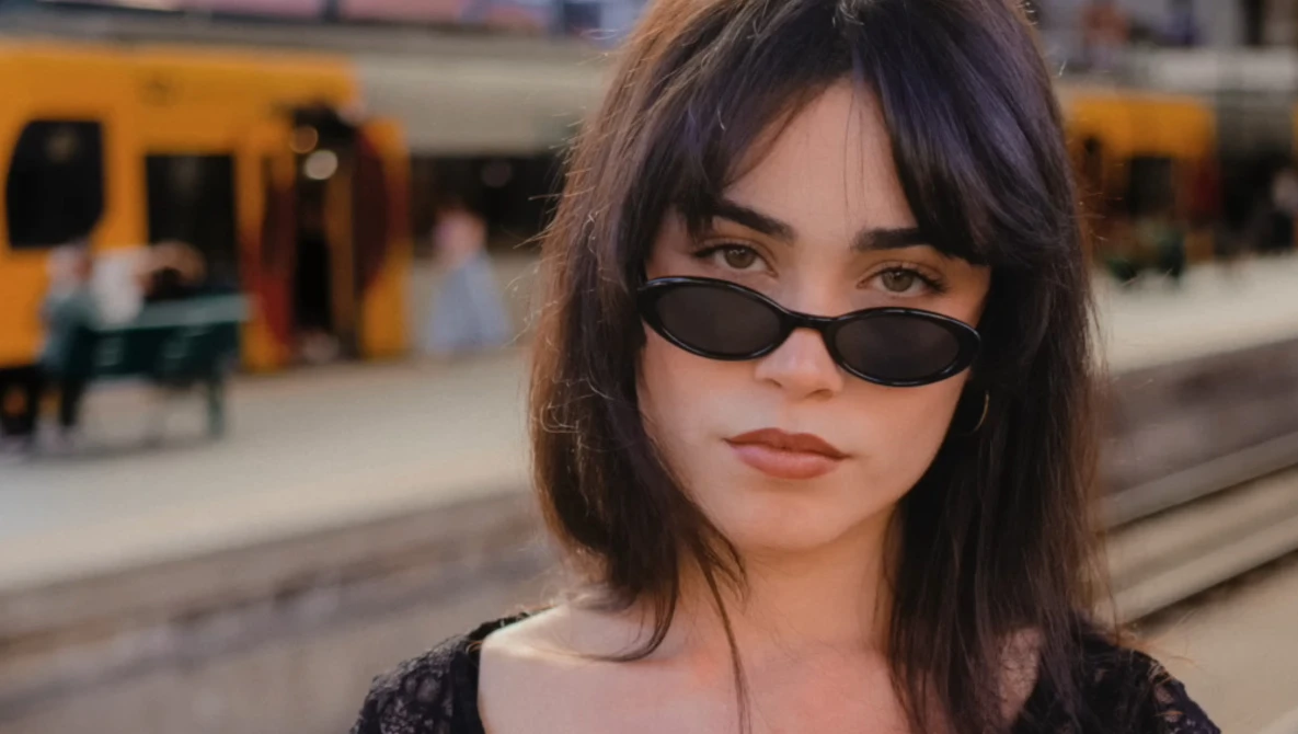 Woman wearing dark oval sunglasses and dark clothing at a train station platform.