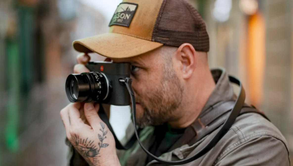 Photographer holding a rangefinder camera up to his eye while wearing a tan and brown trucker cap.