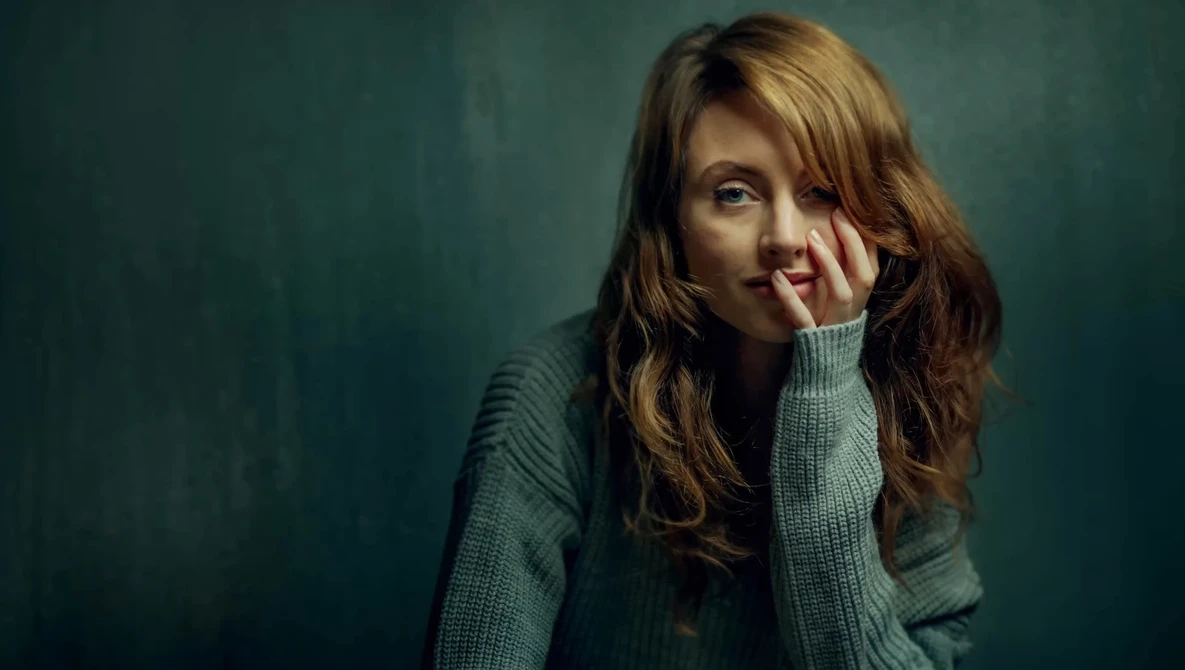 Woman with long reddish-brown hair wearing a gray-green sweater, resting her face against her hand against a teal background.