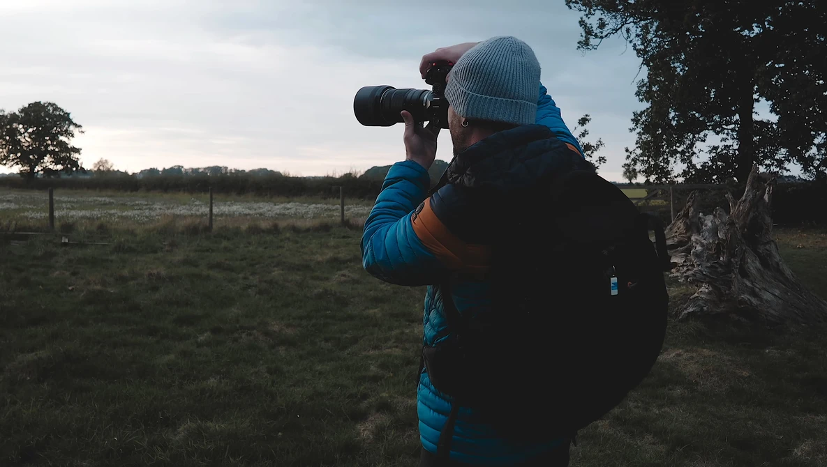 Photographer in blue jacket and beanie using telephoto lens in rural pasture at dusk.