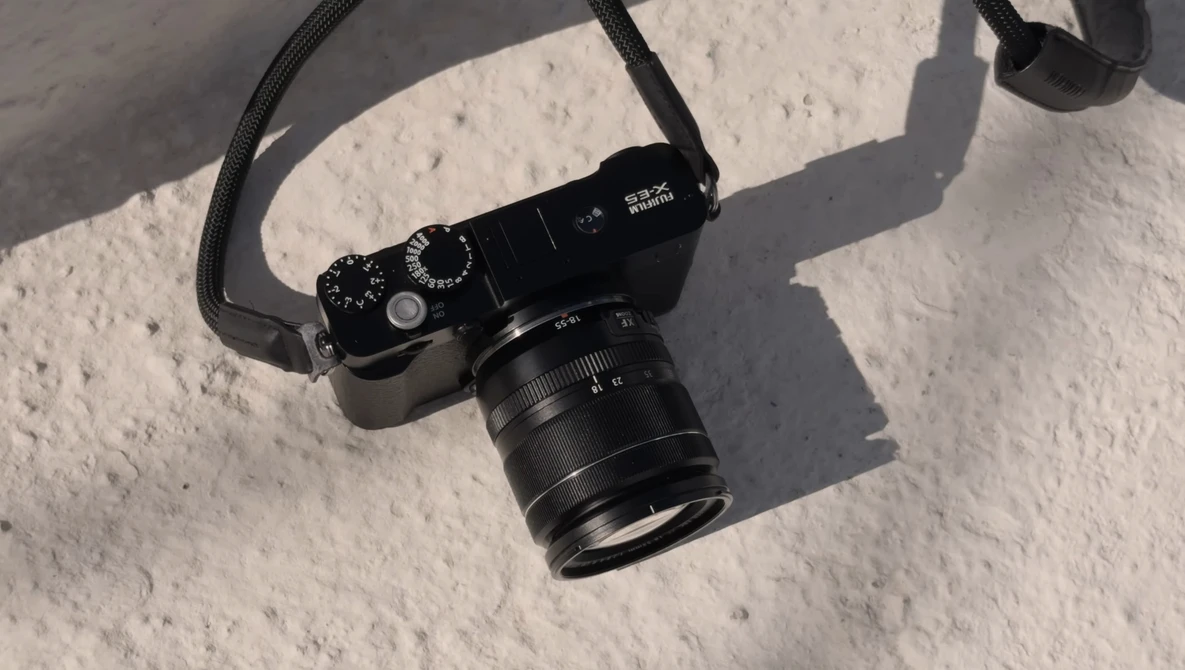 Black rangefinder camera with attached lens on white textured surface, casting sharp shadows in bright sunlight.