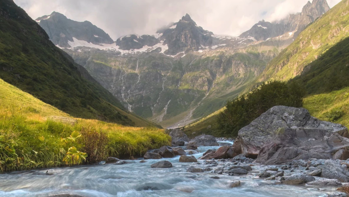 Alpine valley with turquoise glacial stream, green meadows, and snow-capped peaks with waterfalls.