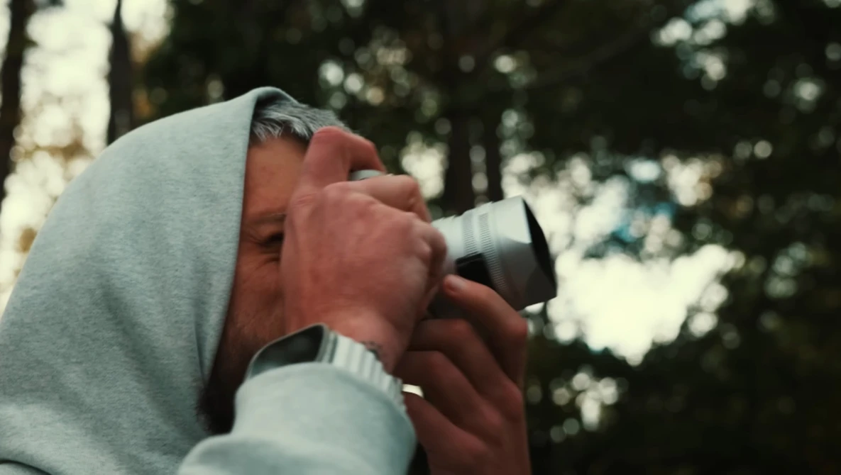 Photographer holding binoculars to eyes while wearing a hooded sweatshirt outdoors.