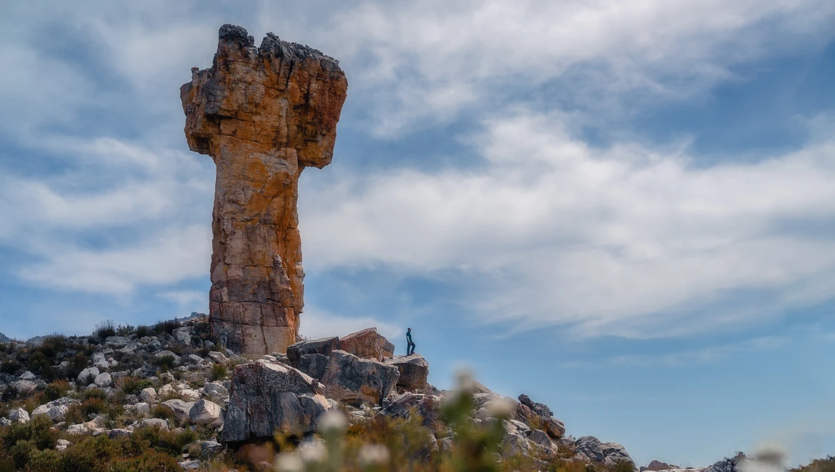 Tall rock tower formation rising from a rocky hillside with a small human figure standing nearby for scale.