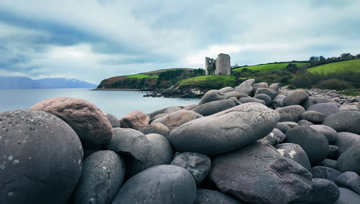 Boulder-strewn beach with castle ruins on distant headland under dramatic cloudy sky.