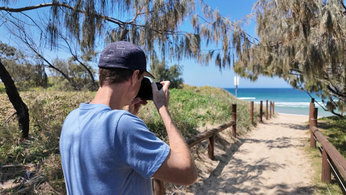 Photographer with camera raised to face, composing a shot down a sandy beach path lined with wooden railings and coastal vegetation.