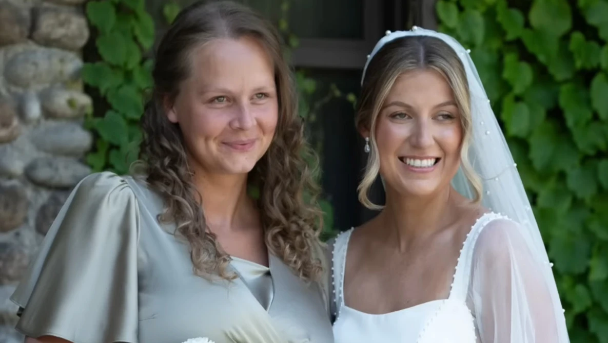 Two women smiling at camera, one wearing a white bridal veil and dress, the other in a light gray sleeve dress.
