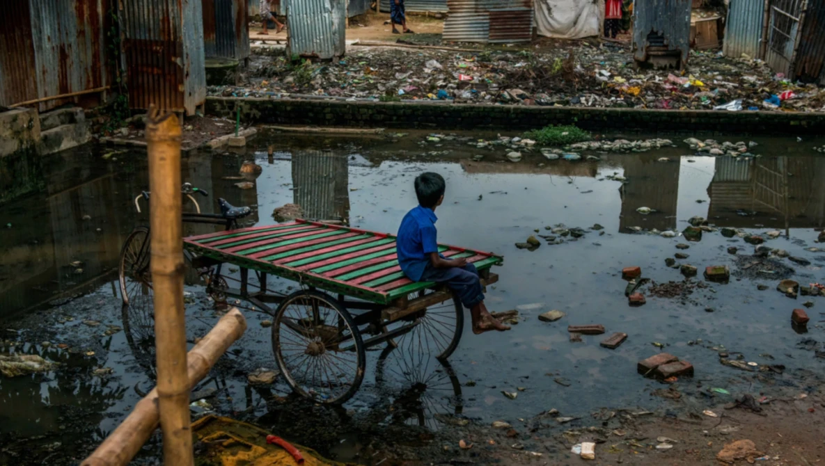 Child in blue shirt sitting on colorful cart in shallow water amid urban debris and waste.