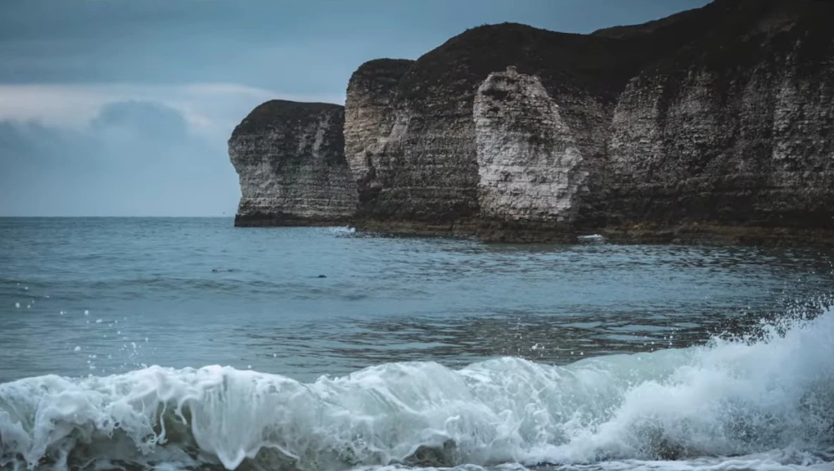 Dramatic limestone sea cliffs rising from foaming ocean waves under a partly cloudy sky.