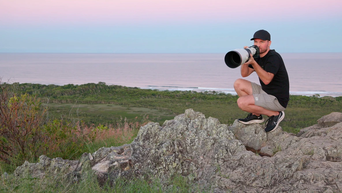 Photographer crouching on rocky outcrop while shooting with telephoto lens toward distant shoreline.