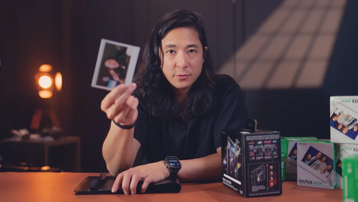 Creator holding instant film photograph at desk surrounded by camera equipment and lighting.