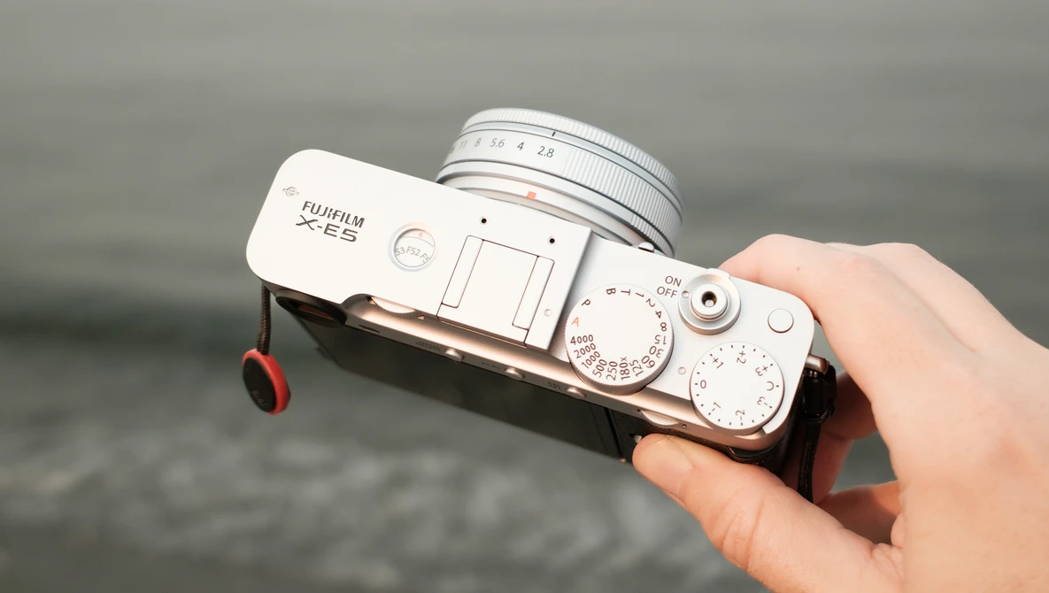 White Fujifilm X-E3 mirrorless camera with attached silver lens held in a hand against a blurred gray water background.
